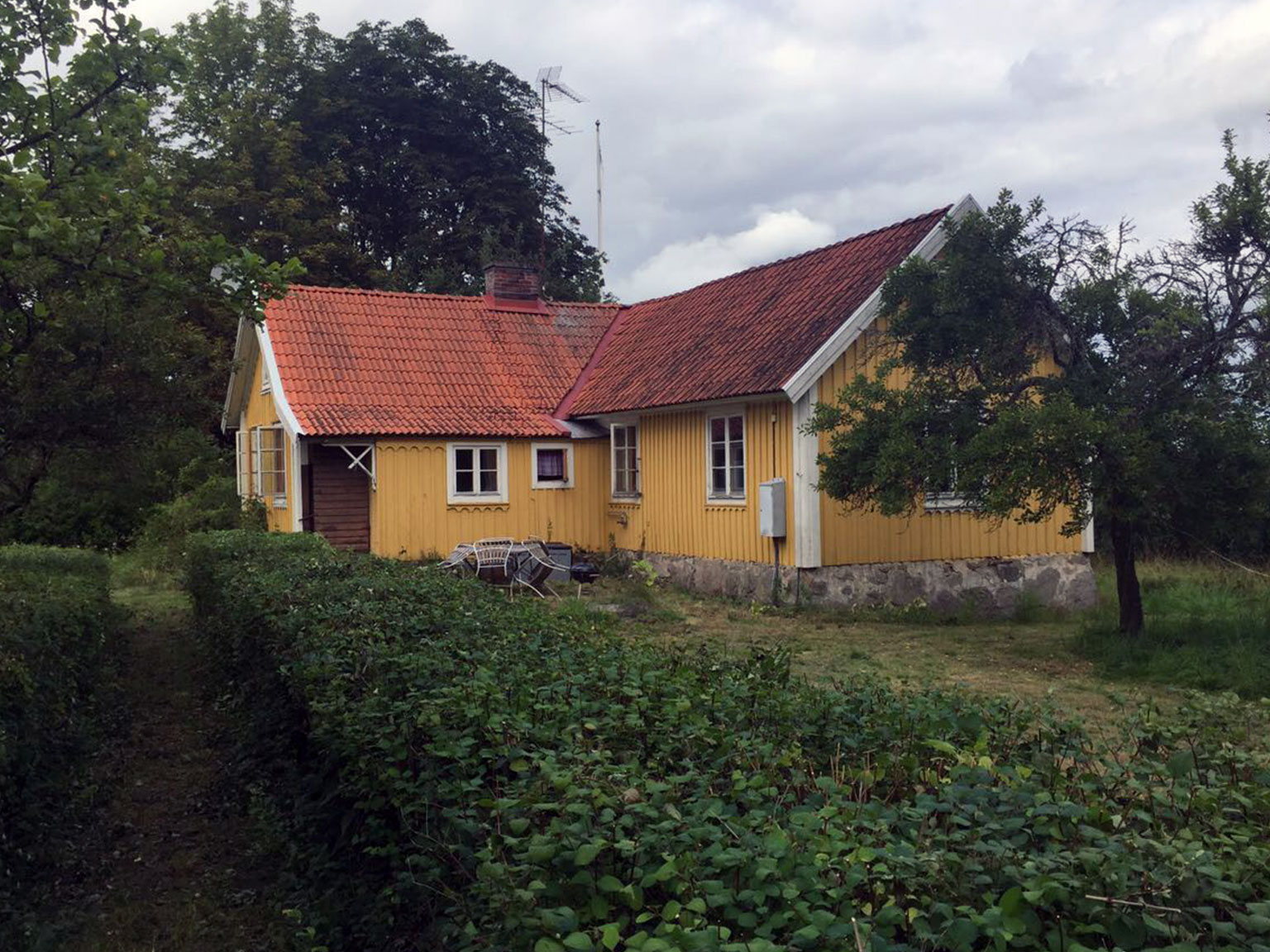 Älteres gelbes Holzhaus in Schweden mit rotem Ziegeldach, Schornstein und verwachsenem Vorgarten, typisches Beispiel für eine renovierungsbedürftige Immobilie beim Hauskauf in Schweden
